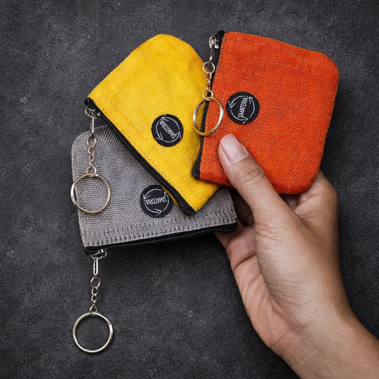 Three Smateria Geek coin purses in yellow, orange, and grey held by a hand against a dark background.