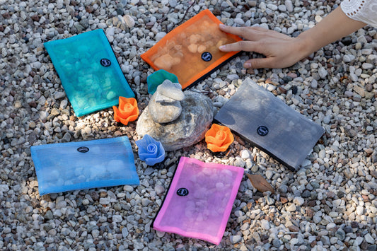 Five Smateria Alpha pouches in signature colours, made from transparent net fabric, laid on a rocky shoreline near the water.
