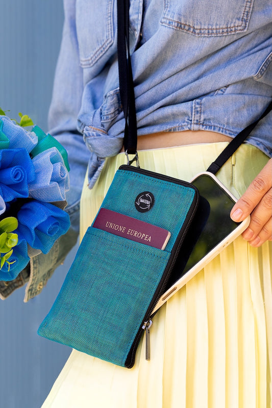 Person holding a Smateria Key crossbody bag with a passport, wearing a blue denim jacket and yellow skirt, against a light blue background.