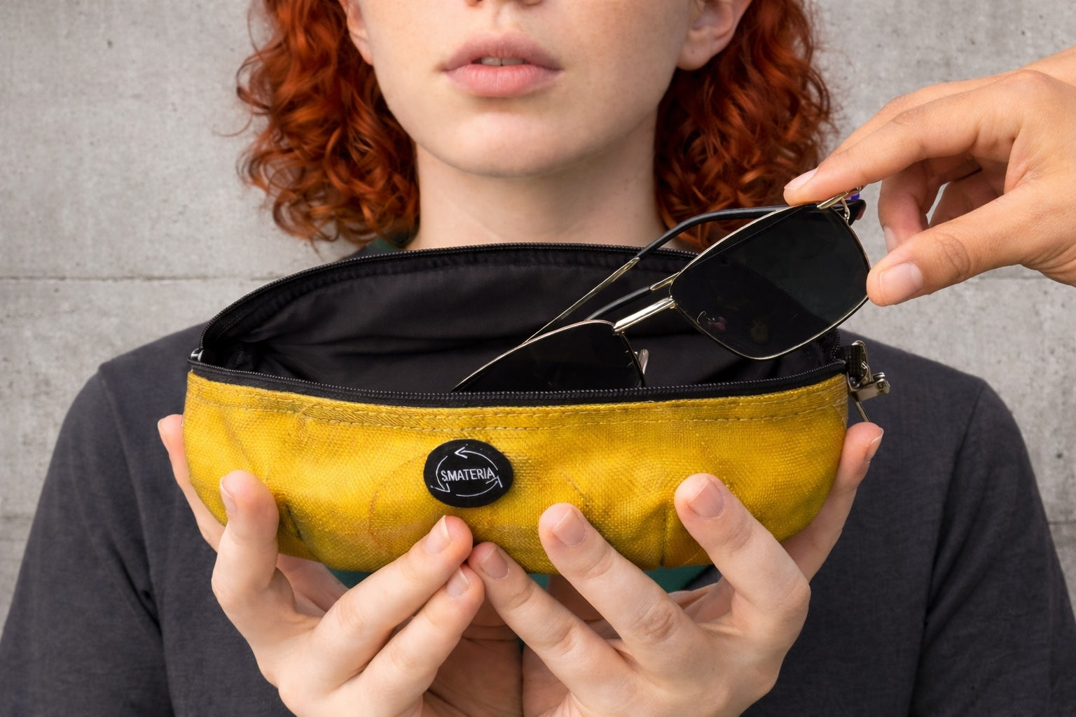 Person holding a Smateria Wink sunglasses case in yellow with sunglasses, against a grey background
