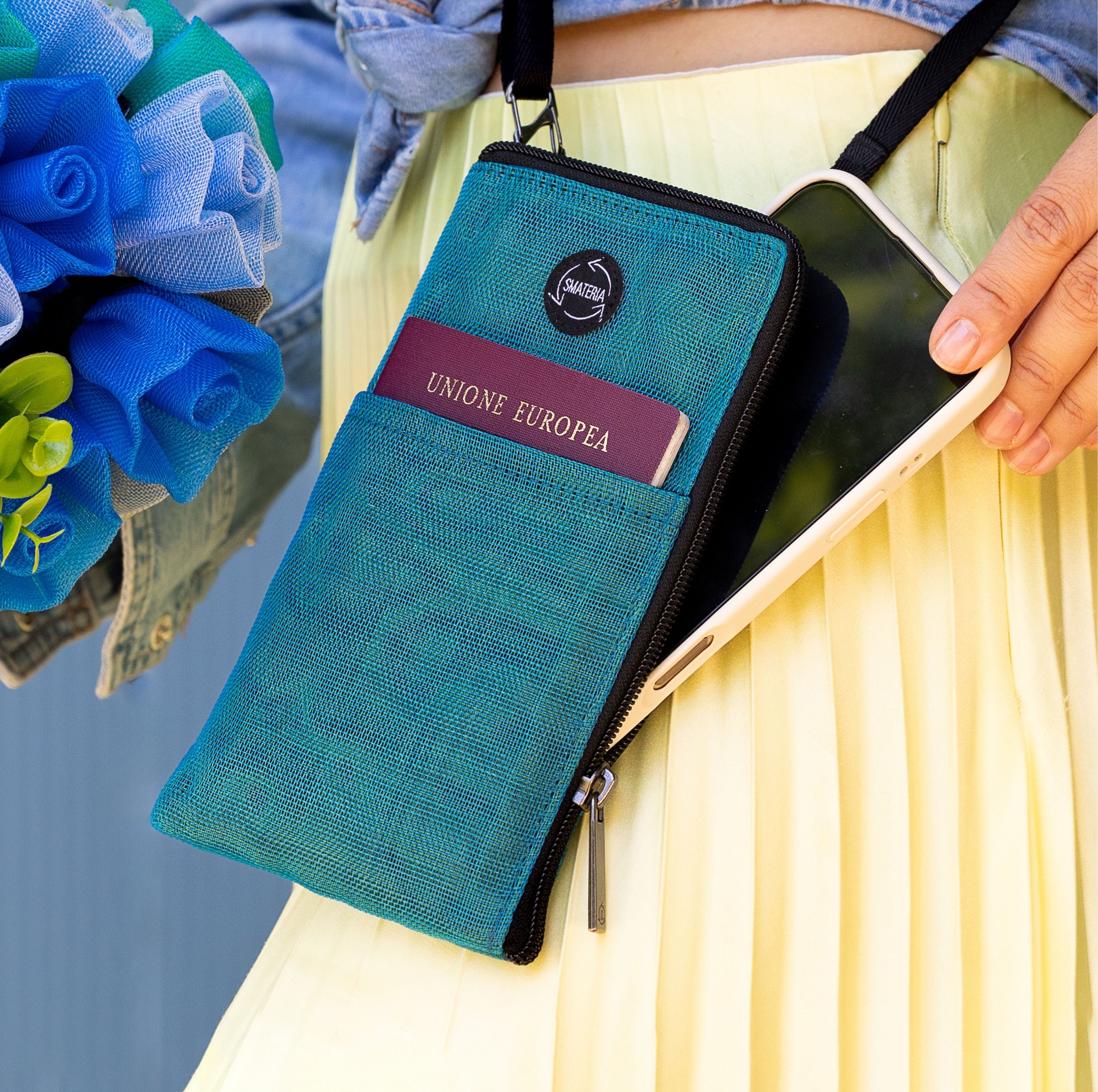 Smateria Key crossbody phone bag with phone and passport held by a person, with flowers in the background