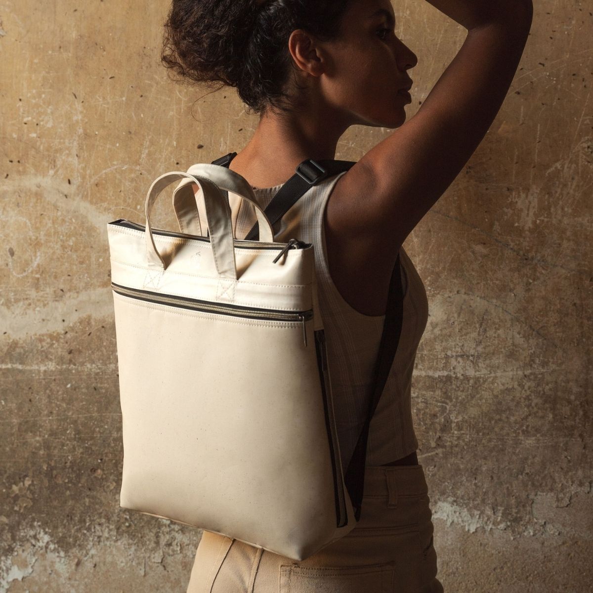 A woman wearing a beige sleeveless top carries a Smateria Momentum natural white backpack with top handles and black straps against a textured concrete wall.