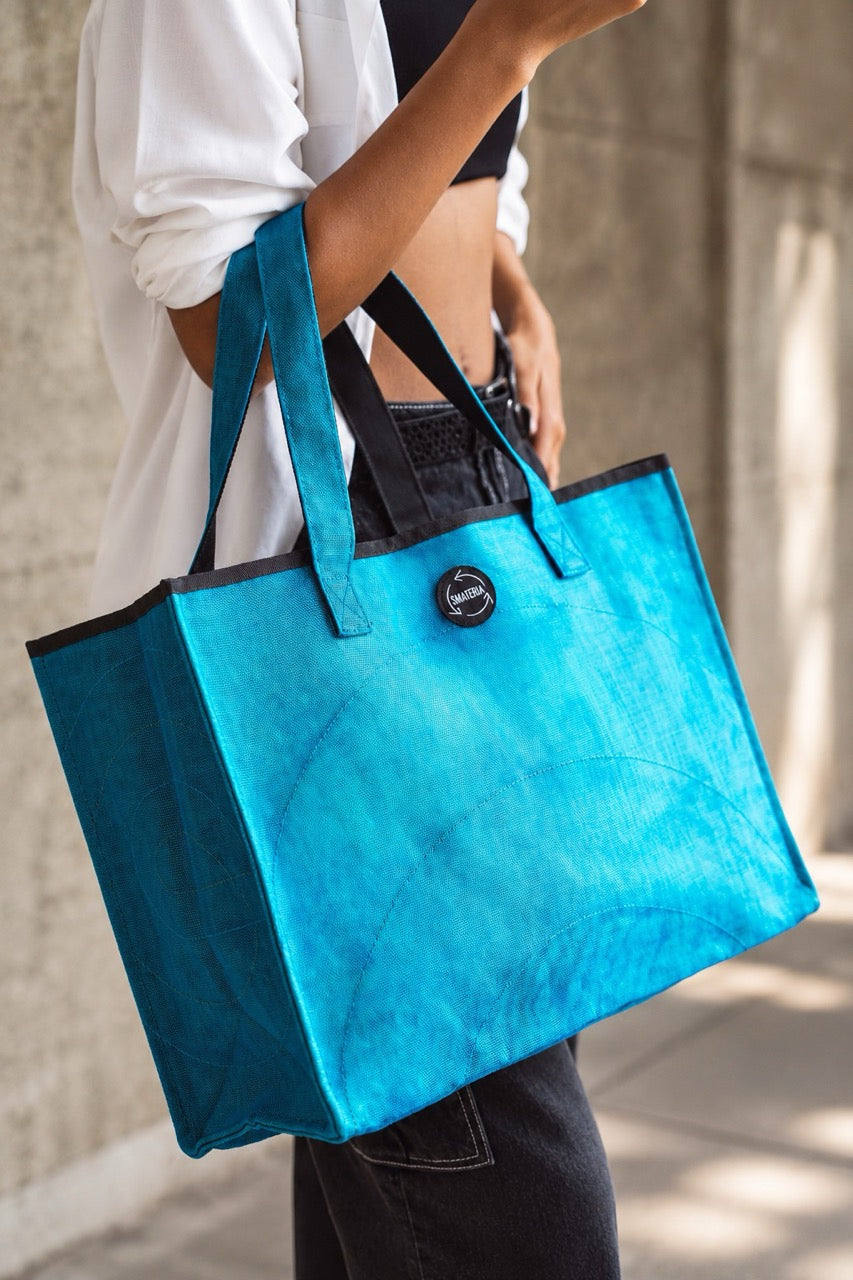 A person carrying a large Smateria Shopper reusable tote in Petrol Blue blue, photographed outdoors against a blurred concrete wall.
